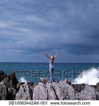 Arquivo de Fotografia - jovem,   mulher, 
  ficar,   pedras, 
  mar,   braços, 
  esticado. fotosearch 
- busca de fotos, 
imagens e clipart