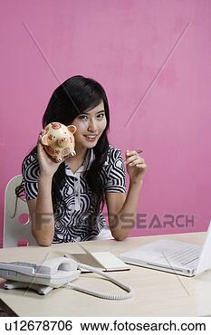 Banco de Imagem - jovem,  mulher, 
 segurando,  porca, 
 poupar,  sentando, 
 escrivaninha. 
fotosearch - busca 
de fotos, imagens 
e clipart