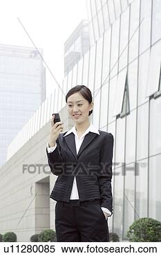 Stock Image - business young 
woman standing, 
holding a mobile 
phone, smiling. 
fotosearch - search 
stock photos, 
pictures, images, 
and photo clipart