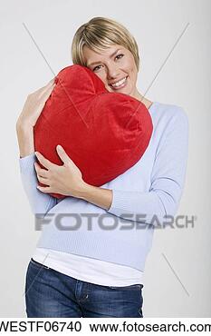Arquivo de Fotografias - jovem,   mulher, 
  segurando,  
 heart-shaped, 
  almofada,   
sorrindo. fotosearch 
- busca de fotos, 
imagens e clipart